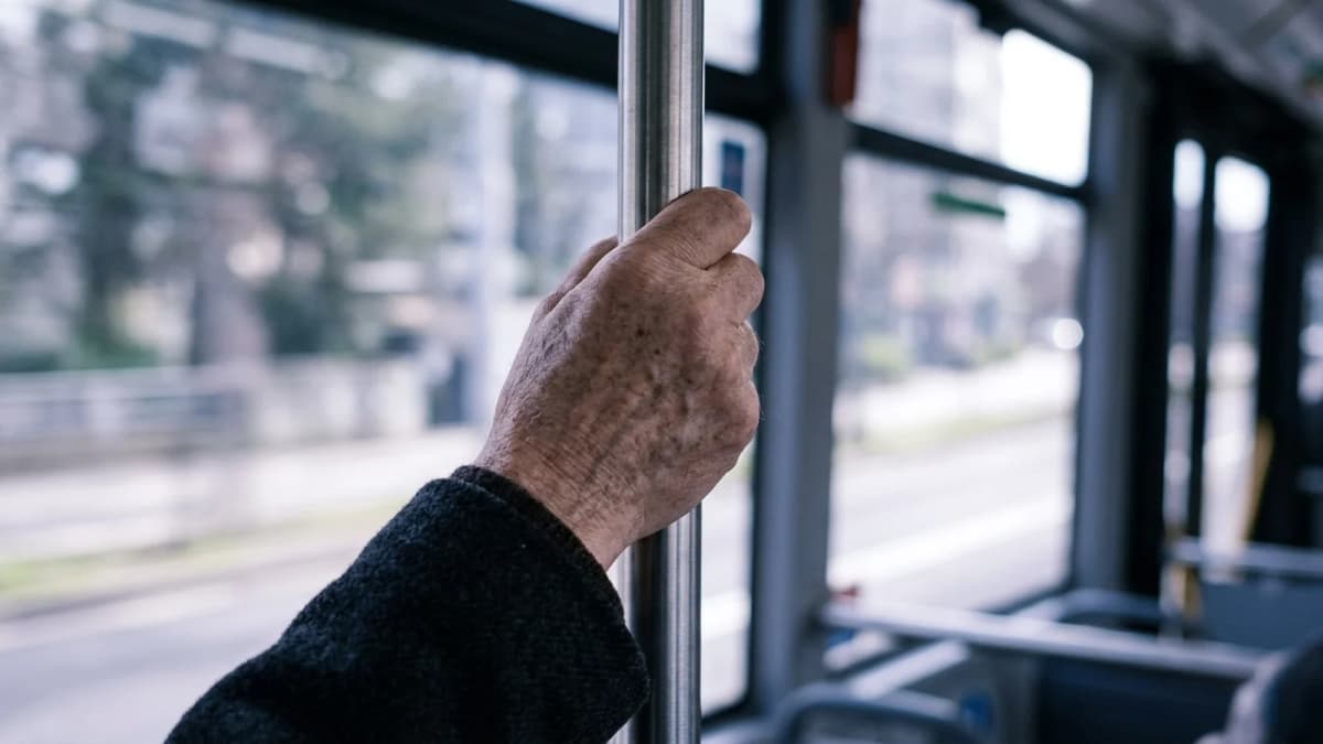 An older hand gripping a metal handrail inside a moving bus