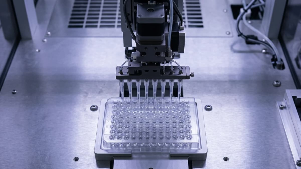 An automated pipetting head above a clear multi-well plate, seen from above under cold light