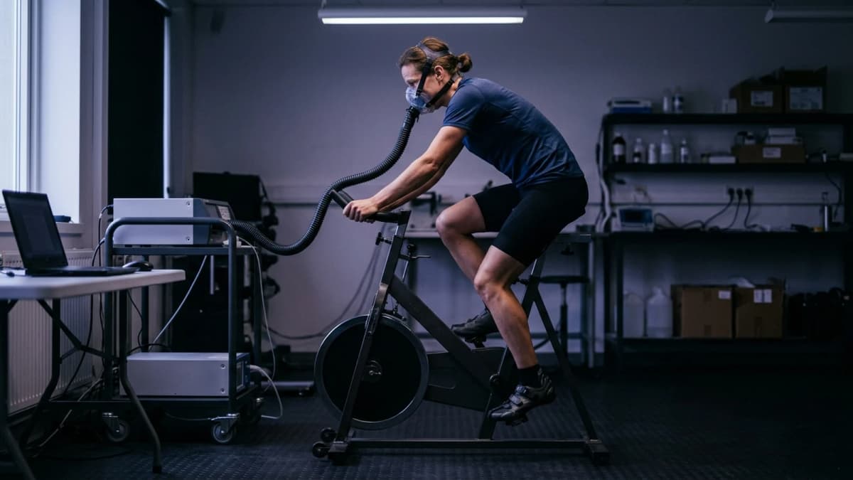 A person on a cycle ergometer wearing a breathing mask with a corrugated hose in a dim exercise lab