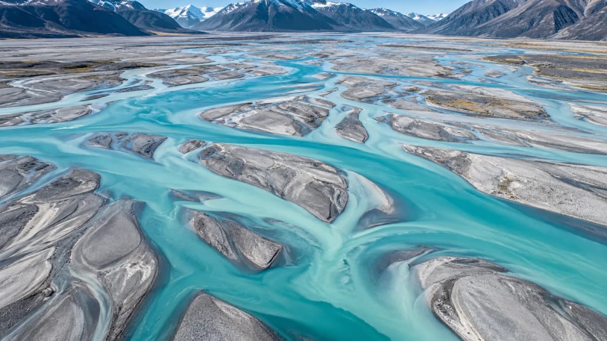 Aerial view of a wide glacial braided river spreading into many turquoise channels across a pale grey gravel plain, snow-capped mountains in the distance, bright midday light